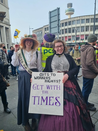 Photo shows two ladies in Edwardian/Victorian costumes with a placard saying "Cuts to workers' rights? Get with the times"