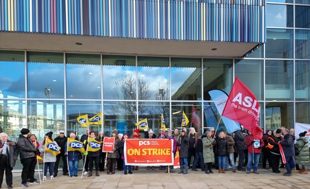 Photo shows long row of pickets outside a modern glass building, with union flags