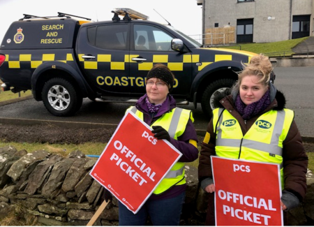 Photo shows two women holding OFFICIAL PICKET placards in front of a black and yellow Coastguard vehicle