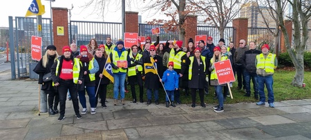 Image shows a group of about 25 PCS members with placards, and high vis vests by the gates of a building.