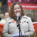 Fran is standing behind a microphone wearing a coat with a PCS on Strike sticker