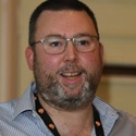 Ian has short dark hair and a grey beard, and wears glasses and an open-necked shirt