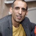 Zulf has short, dark hair and is wearing a yellow tshirt and grey jacket