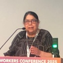 Tracey has long dark hair in a ponytail, wears glasses and is speaking at a lecturn