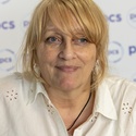 Angela has blonde hair and is wearing a white shirt, standing in front of a PCS backdrop