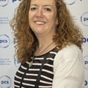 Fran has long curly hair and is sitting in front of a PCS backdrop, smiling at the camera