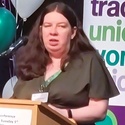 Liz has long brown hair. She is standing behind a lecturn, making a speech.