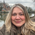 Fern has shoulder-length, fair hair. She is standing by a river with a church in the background.