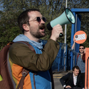 The photo shows a man wearing sunglasses and holding a megaphone with a 4-day week placard in the background.