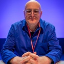 The photo by Andy Aitchison shows PCS Assistant General Secretary John Moloney seated wearing glasses a blue shirt and a red lanyard at PCS Annual Delegate Conference in Brighton in May 2026